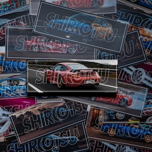 May include: A pile of rectangular stickers depicting sports cars in motion. Each sticker has a black border and the word "SHIROTSU" in a white, stylized font. The cars are various colours, including red, blue, and purple.