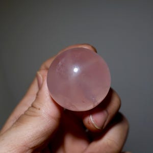 May include: A polished rose quartz crystal sphere, held in a hand. The ball is a soft pink colour, with a translucent appearance. The background is a plain grey, highlighting the crystal's form and colour.