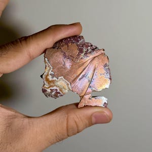 Könnte beinhalten: Ein flaches, unregelmäßiges Mineralexemplar mit einer metallisch kupferfarbenen Oberfläche und einer strahlenförmigen, fächerartigen Textur. Die Kanten zeigen raue, erdbraune und weiße kristalline Schichten, die einen Kontrast zur glatten, reflektierenden Mitte bilden.