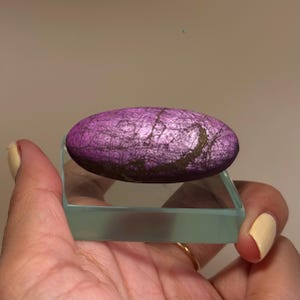 May include: A purple, oval-shaped stone with dark veining rests on a clear, rectangular glass base. The stone is held in a hand with a yellow painted fingernail. The stone has a textured surface.