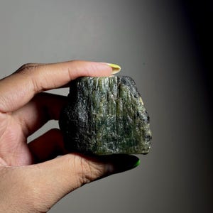 May include: A raw, unpolished mineral specimen featuring a deep forest green color with vertical striations and a rough, textured surface. The stone displays varying shades of dark olive and black, with a semi-translucent quality in certain areas.