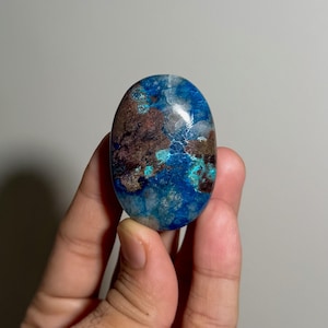 以下が含まれることがあります： 深い青、鮮やかなターコイズ、そして暗い茶色の土のようなインクルージョンが混ざり合った、楕円形の磨かれた宝石。石の表面は滑らかで光沢があり、全体に大理石のような模様と自然な色の変化が見られます。