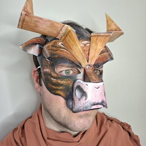 Könnte beinhalten: Eine Papier-Bullenmaske in Braun- und Beigetönen mit großen Hörnern und einem detaillierten Gesicht. Die Maske bedeckt das obere Gesicht, wobei die Augen des Trägers sichtbar sind. Die Maske wird über einem braunen Kleidungsstück getragen.