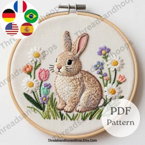 May include: Embroidered bunny in a wooden hoop. The bunny is light brown with pink ears, surrounded by colorful flowers and green grass. The text "PDF Pattern" is visible.