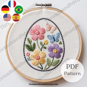 Puede incluir: Diseño floral bordado en un aro de madera. El diseño presenta flores rosas, amarillas y moradas, hojas verdes y dos mariposas. El bordado está sobre un fondo blanco. El texto "PDF Pattern" es visible.