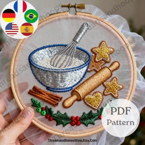 May include: Embroidered hoop art featuring baking elements. A silver whisk sits in a blue-rimmed bowl filled with flour. A rolling pin, gingerbread cookies, and cinnamon sticks are also depicted. The design includes a holly and berry border. The text "PDF Pattern" is visible.