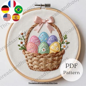 以下が含まれることがあります： カラフルな刺繍のイースターエッグが入った織りバスケットが特徴の刺繍枠。バスケットにはピンクのリボンが付いており、刺繍の花で飾られています。「PDF Pattern」の文字が見えます。ThreadsandhoopsShop.Etsy.comのウェブサイトも表示されています。