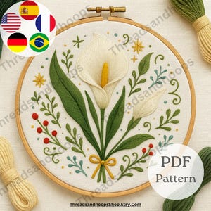 May include: An embroidery hoop with a white calla lily design. The floral design includes green leaves, red berries, and yellow accents. The text "PDF Pattern" is visible on the lower right. The website ThreadsandhoopsShop.Etsy.com is also visible.