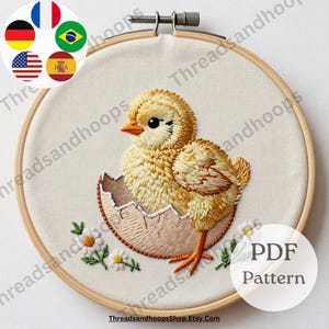 Puede incluir: Pollito bordado saliendo de un huevo, en un bastidor de bordado de madera. El pollito es amarillo con pico y patas naranjas. La cáscara del huevo es marrón claro, y pequeñas flores blancas con centros amarillos están bordadas debajo. El texto "PDF Pattern" es visible.