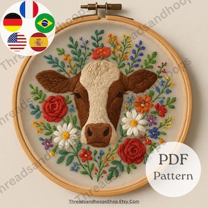 May include: Embroidered cow face with floral accents in a wooden hoop. The cow has brown and white fur, surrounded by red roses, daisies, and other colorful flowers. The text "PDF Pattern" is visible on the right side of the hoop.