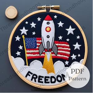May include: Embroidered hoop art featuring a rocket ship, American flag, and stars against a navy blue background. The word "FREEDOM" is stitched in white. The design is framed in a wooden hoop.
