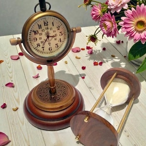 Peut inclure: Un ensemble horloge et sablier de style ancien. L'horloge a un cadran rond avec des chiffres romains et une finition bronze. Le sablier est en bois et en verre. Le fond comprend des fleurs roses et des pétales épars.