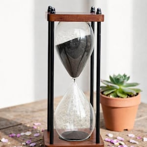 Könnte beinhalten: Sanduhr mit Holzrahmen und schwarzem Sand. Die klare Glaskugel wird von schwarzen Metallstäben getragen. Eine kleine Sukkulente in einem Terrakottatopf und verstreute Blütenblätter befinden sich im Hintergrund.