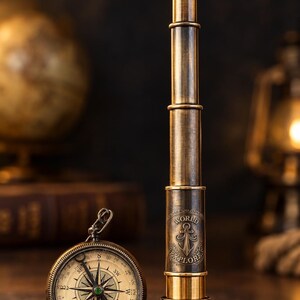 Peut inclure: Une longue-vue nautique en laiton et une boussole de poche ronde. La longue-vue présente une ancre gravée, le texte "WORLD EXPLORE" et "ADVENTURE AWAITS". La boussole possède un cadran circulaire avec les points cardinaux et une aiguille.