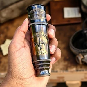 May include: A vintage brass telescope with the text "DOLLOND LONDON 1920" and a fleur-de-lis emblem. The telescope has a dark metallic finish and is held in a hand.