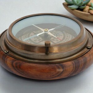 May include: A vintage compass with a brass bezel and a wooden base. The compass face is visible through a glass cover, with directional markings and a needle. The wooden base has a rich, dark brown grain. A decorative item.