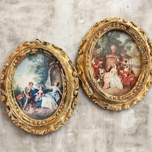 May include: Two oval wall art pieces featuring ornate, textured gold frames. The left piece depicts a group of figures in period clothing in a garden setting. The right piece shows a gathering of people in colorful attire near a stone pedestal.