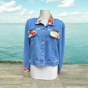Könnte beinhalten: Hellblaue Jeansjacke mit einzigartigem Design. Der Kragen hat einen Zebrastreifen und florale Akzente. Die Taschen und Manschetten haben florale Muster. Dekorative Perlen schmücken die Taschen. Die Jacke hat vier Knöpfe vorne.