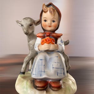 May include: A ceramic figurine of a girl in a bonnet and dress, holding orange flowers. A gray lamb stands behind her. The figurine has a painted design with brown shoes and is on a green base.