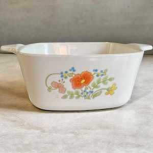 May include: A white rectangular ceramic baking dish with integrated side handles. The exterior features a central floral decal with a large orange poppy, small blue forget-me-nots, green leaves, and yellow accents on a smooth, glossy white surface.