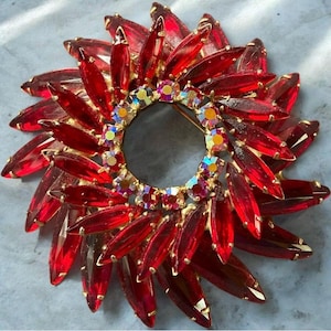 May include: A circular brooch featuring multiple layers of pointed, marquise-cut red glass stones arranged in a sunburst pattern. The center of the brooch is accented with a ring of smaller, iridescent, multi-colored round rhinestones set in gold.