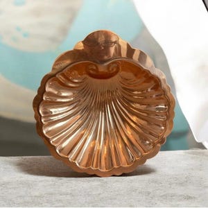 May include: A copper-colored, shell-shaped decorative dish. The dish has a scalloped edge and a detailed, ridged interior. The metallic surface reflects light, creating highlights and shadows. The dish is likely used for decorative purposes or serving small items.