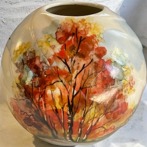 May include: A round ceramic vase with a hand-painted autumn tree design. The vase features a cream-colored base with a colorful depiction of a tree with red, orange, and yellow leaves. The tree's dark branches contrast against the lighter background.