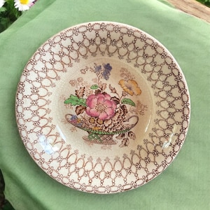 May include: A vintage ceramic plate with a floral design. The plate features a central bouquet of pink, yellow, and blue flowers in a decorative basket. The rim has a brown, openwork pattern. The plate is set on a green cloth.