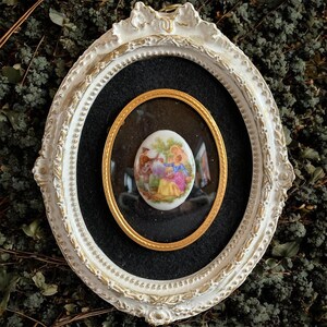 May include: An oval framed miniature painting. The white frame has gold accents and a decorative design. Inside, a gold-rimmed oval surrounds a black background, showcasing a smaller oval painting with a pastoral scene in vibrant colors.