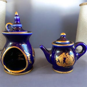 May include: Two-piece cobalt blue porcelain set with gold trim. The set includes a small teapot with a lid and a decorative container with an opening. The teapot features a gold relief design of figures. The set is likely vintage.