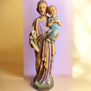 May include: A religious statue depicting a man holding a child. The man wears a robe with purple and gold accents, and holds flowers. The child is dressed in blue. The statue has a detailed, painted finish.