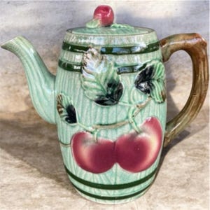 May include: A ceramic teapot with a light green crackle glaze finish, featuring a barrel-like texture with dark green horizontal bands. The body is decorated with embossed red cherries and green leaves, a brown branch-shaped handle, and a lid with a small red cherry finial.