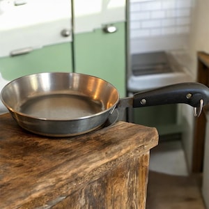 Puede incluir: Una pequeña sartén de acero inoxidable con un mango negro. La sartén está sobre una superficie de madera. El mango tiene un lazo para colgar. La sartén está vacía y parece sin usar.