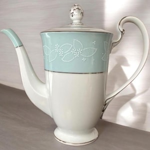 Może przedstawiać: Wazon do kawy z porcelany w stylu vintage z jasnoniebieskim paskiem z kwiatowym wzorem. Dzbanek ma białą podstawę, zakrzywiony uchwyt i dzióbek. Pokrywka zwieńczona jest ozdobnym guzikiem i ozdobiona złotymi akcentami. Klasyczny element do serwowania kawy lub herbaty.