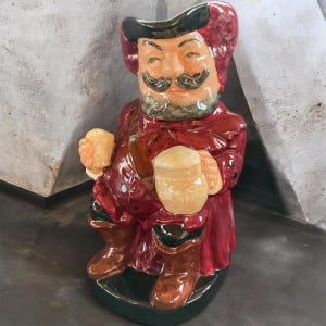May include: A ceramic figurine of a man in a red coat and hat, holding two mugs of beer. The figurine has a mustache and beard, and is wearing brown boots. The base is dark green.