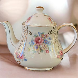 May include: A cream-colored porcelain teapot featuring a delicate floral bouquet of pink, yellow, and blue flowers. The teapot includes a matching lid, a curved handle, and a spout, all accented with fine gold trim and a subtle gold lattice pattern.