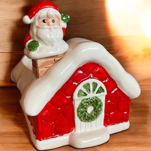 May include: A ceramic Christmas cookie jar shaped like a red house with white snow-covered roof and a Santa Claus figure on top. The house has a white door with a green wreath and a chimney. The Santa figure is wearing a red hat and suit.
