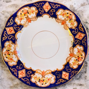 May include: A decorative plate with a white center and a deep blue rim. The rim features gold filigree and orange floral designs. The plate has a scalloped edge and a gold inner ring. A classic piece for serving or display.