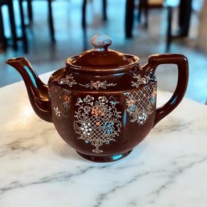 May include: A brown teapot with a floral design and gold accents. The teapot has a lid and a spout. It is sitting on a white marble surface.