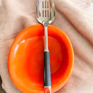 Könnte beinhalten: Ein Schöpflöffel mit schwarzem und orangefarbenem Griff liegt auf einer orangefarbenen Schüssel. Der Löffel besteht aus glänzendem Metall und hat eine Reihe rechteckiger Schlitze. Die Schüssel ist rund und hat eine glänzende Oberfläche. Löffel und Schüssel liegen auf einem beigen Tuch.