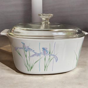 May include: An oval white casserole dish with integrated side handles and a clear glass lid featuring a knob handle. The exterior of the dish is decorated with a pattern of light purple iris flowers, green stems, and thin vertical blue and purple lines.