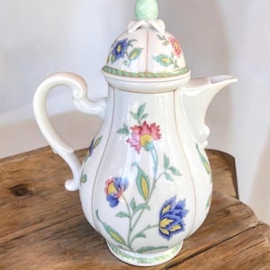 Może przedstawiać: Biały porcelanowy czajnik z kwiatowym wzorem. Czajnik ma pokrywkę z zielonym uchwytem i rączką. Czajnik jest ozdobiony różowymi, żółtymi, niebieskimi i zielonymi kwiatami.