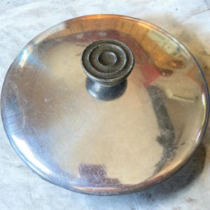 Op de afbeelding: Een rond deksel van gepolijst roestvrij staal met een reflecterend oppervlak. Het deksel is voorzien van een centraal geplaatste, donkergekleurde, ronde knop met een getrapt, geribbeld ontwerp. Het metalen oppervlak vertoont lichte krasjes.