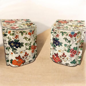 Può includere: Due scatole decorative con motivi floreali. Le scatole sono bianche con un motivo ripetuto di fiori rossi, arancioni, blu e viola e foglie verdi. Le scatole hanno una forma curva e un coperchio.