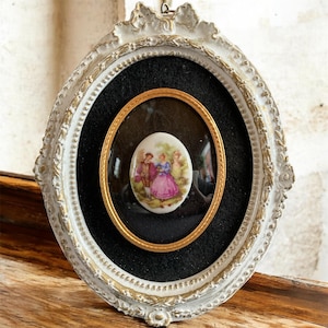May include: An oval-shaped, antique-style frame with a white and gold finish. Inside, a black background surrounds a smaller gold frame, which holds a porcelain cameo with a painted scene of figures in period clothing. The outer frame has ornate detailing.