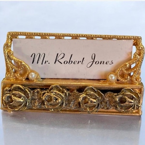May include: A gold-toned metal name card holder featuring a rectangular frame with ornate scrollwork corners, two small white faux pearls, and a base decorated with four sculpted metal roses. A white card with the text Mr. Robert Jones is inserted.