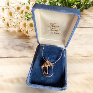 May include: A gold-colored umbrella pendant necklace displayed in a blue velvet jewelry box. The box is open, revealing the necklace against a blue interior. The box is labeled "First Love by LANG".