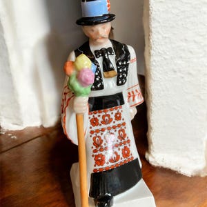 May include: A ceramic figurine of a man in traditional attire. The figure wears a black top hat with a blue band, a black vest with white polka dots, and a white shirt with red floral embroidery. He holds a staff topped with colorful spheres.
