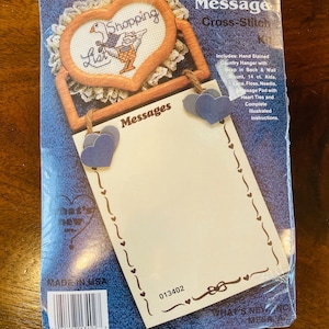 May include: A Country Message Cross-Stitch kit. The kit includes a heart-shaped cross-stitch design with a goose and shopping list, a wooden frame, and a notepad with heart-shaped accents. The package is sealed in plastic.