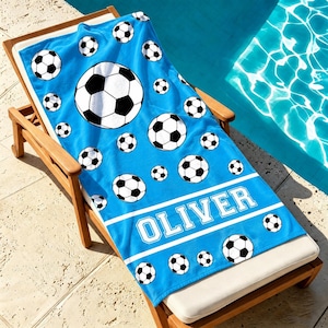 May include: A bright blue beach towel featuring a repeating pattern of black and white soccer balls of various sizes. The towel includes a horizontal white band with the name OLIVER printed in a bold, block-style white font across the lower section.